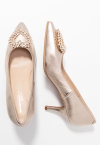 Metallic gold high-heeled shoes with a pointed toe and a jeweled accent in a heart shape on the front, featuring a smooth texture.
