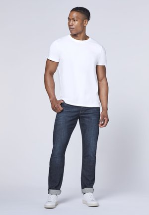 Weißes T-Shirt mit Rundhalsausschnitt, kombiniert mit dunkelblauen, geraden Jeans mit umgeschlagenen Säumen und weißen Sneakers. Glattes Material mit minimalen Details.