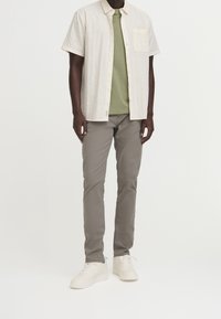 Man som bär en ljusbeige kortärmad skjorta över en olivgrön T-shirt, grå slim-fit byxor och vita sneakers, står framför en vit bakgrund.