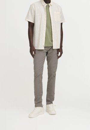 Uomo che indossa una camicia a maniche corte beige chiaro con bottoni sopra una maglietta verde oliva, pantaloni grigi slim-fit e sneakers bianche, in piedi su uno sfondo bianco.