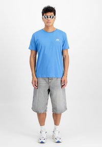 Zils kokvilnas T-krekls ar apaļu kaklu un logotipa detaļu. Valkāts ar pelēkiem džinsu šortiem līdz ceļgaliem un baltām sporta apaviem. Aksesuāri ietver saulesbrilles.