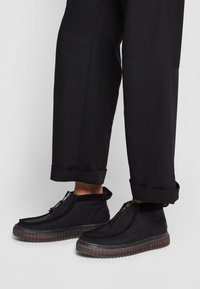 Personne portant un pantalon noir et des chaussures noires montant jusqu'aux chevilles avec des semelles épaisses translucides et des fermetures éclair à l'avant.