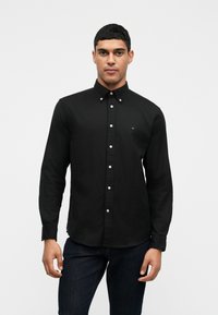 Čierna polokošeľa s dlhými rukávmi a golierom na gombíky, s bielymi gombíkmi a malým logom pri hrudníku. Hladká textúra látky.