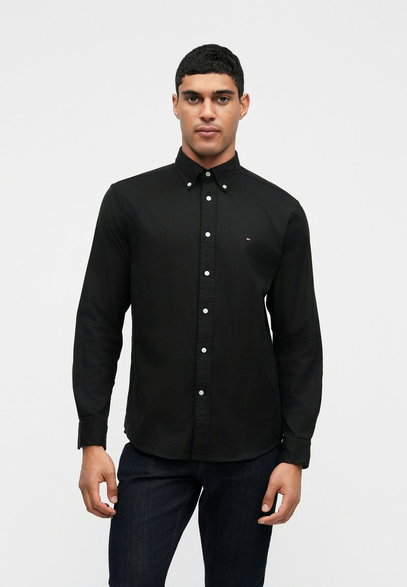 Čierna polokošeľa s dlhými rukávmi a golierom na gombíky, s bielymi gombíkmi a malým logom pri hrudníku. Hladká textúra látky.