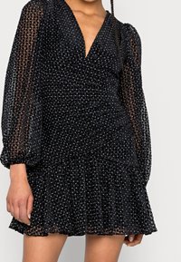 Robe mini noire avec de longues manches transparentes, décolleté en V profond, tissu texturé avec de petits pois et ourlet à volants.