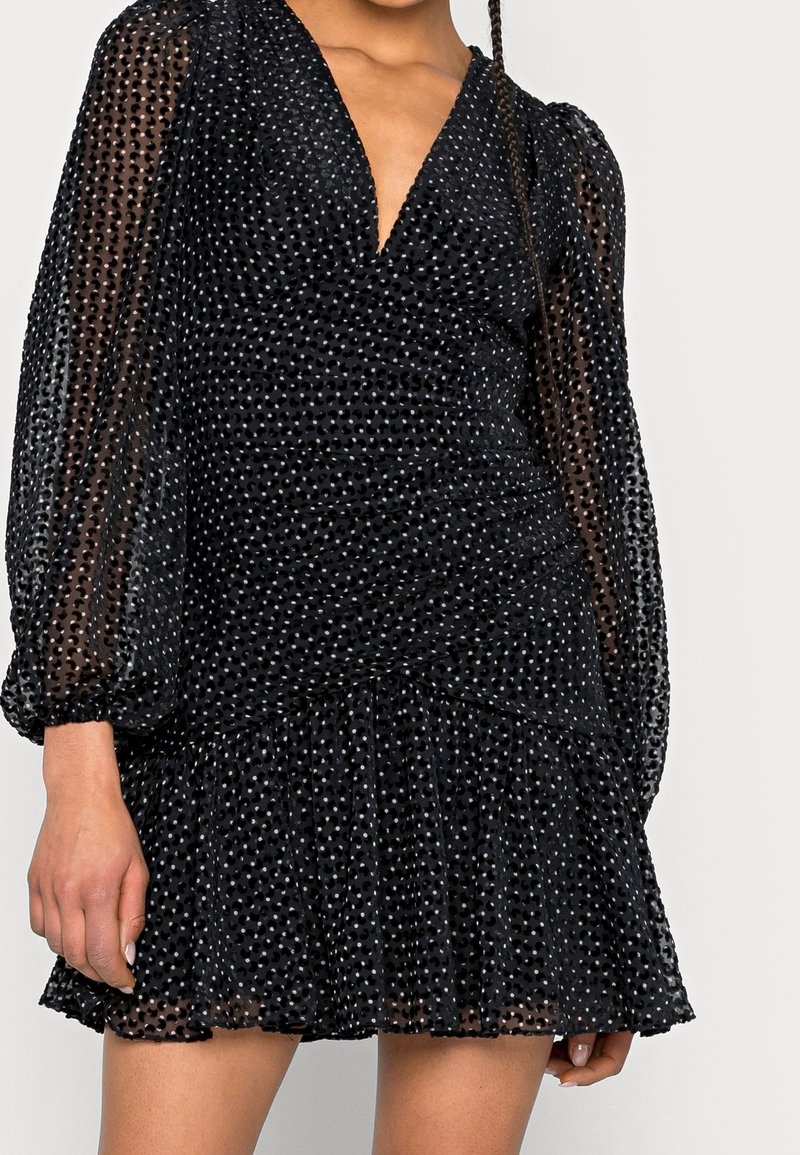 Robe mini noire avec de longues manches transparentes, décolleté en V profond, tissu texturé avec de petits pois et ourlet à volants.