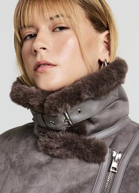 Veste avec un col en fausse fourrure grise et une fermeture éclair brillante, présentant une texture douce, des matériaux contrastés et un design structuré.