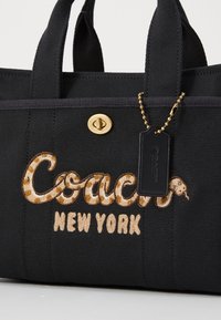 Sac cabas en toile noire avec le mot "Coach" brodé dans un motif serpent. Il présente des ferrures dorées et une étiquette noire suspendue à une chaîne.
