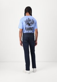 Light blue t-shirt with a printed graphic on the back; dark blue jeans; white sneakers; medium build, standing, facing away from the camera.
