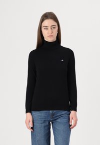 Pulover negru cu gât, confecționat dintr-un material moale; design ajustat, cu mâneci lungi și un mic accent cu logo pe piept. Purtați cu blugi albaștri.