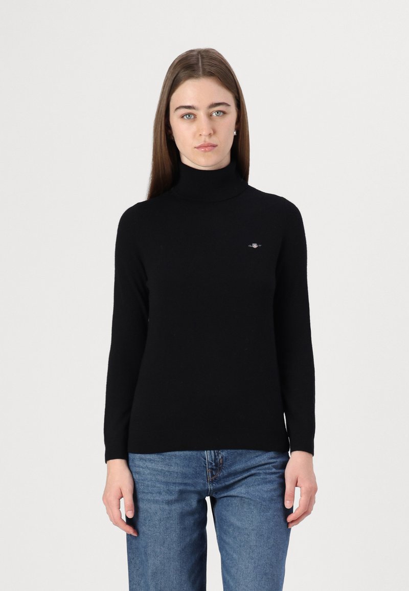 Pulover negru cu gât, confecționat dintr-un material moale; design ajustat, cu mâneci lungi și un mic accent cu logo pe piept. Purtați cu blugi albaștri.