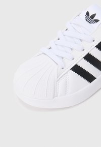 Άσπρο αθλητικό παπούτσι Adidas με μαύρες ρίγες, σχέδιο με μύτη σε σχήμα καβουκιού και άσπρα κορδόνια σε ουδέτερο φωτεινό φόντο.
