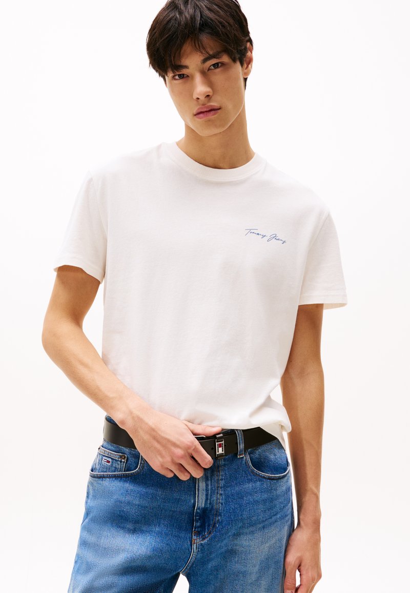 Fehér pamut póló kerek nyakkivágással, bal mellen kék "Tommy Jeans" hímzéssel, kék farmernadrággal és fekete övvel párosítva.