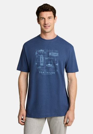 Man draagt een blauw grafisch T-shirt met boot- en haaienontwerpen en lichtgrijze broek, staand tegen een effen witte achtergrond.