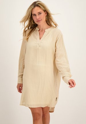 Blonde vrouw draagt een beige, lange mouwen, knie-lange jurk met dunne verticale strepen en gouden creolen.