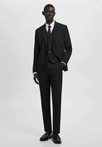 Schwarzer Anzug mit einem Reverskragen, drei Knopf Weste, weißem Hemd und schwarzer Krawatte. Polierte schwarze Schuhe. Maßgeschneiderte Passform und glatte Textur.