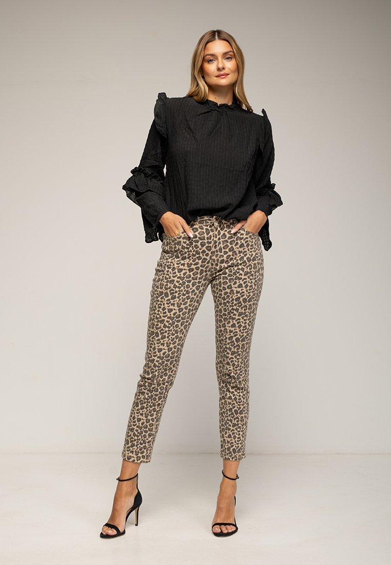 Frau steht mit den Händen in den Taschen, trägt eine schwarze Rüschenbluse, eine Hose mit Leopardenmuster und schwarze High-Heels vor einem schlichten Hintergrund.