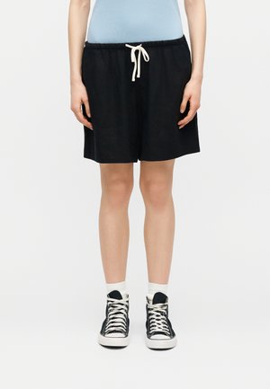 Άτομο που φοράει μαύρο σορτς με κορδόνι, ανοιχτό μπλε μπλουζάκι, λευκές κάλτσες και μαύρα sneakers ψηλού τύπου, στέκεται μπροστά σε λευκό φόντο.