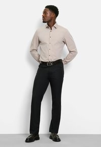 Hellbraunes Hemd mit Knöpfen und vertikalen Streifen, kombiniert mit schwarzen Hosen und einem dunklen Gürtel. Die Schuhe sind aus poliertem schwarzem Leder.