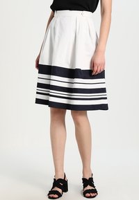 Une jupe plissée à mi-longueur en coton blanc avec un ourlet bleu marine présentant des rayures horizontales. Associée à des sandales à lanières noires.