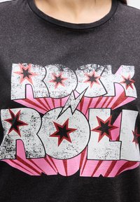 Musta paita, jossa on rohkeat valkoiset ja vaaleanpunaiset "ROCK ROLL" -tekstit, punaiset ja mustat tähtikuvioinnit sekä keskellä salaman kuva.