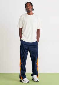 Hvid kortærmet t-shirt parret med marineblå træningsbukser med orange sidelister og accenter. Skoene er hvide sportssneakers.