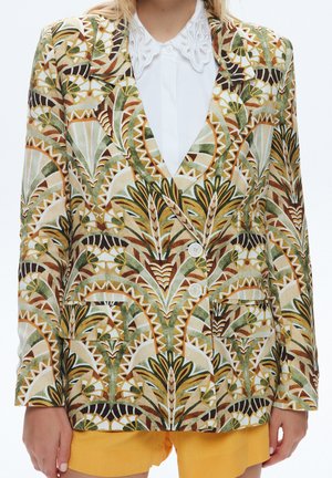 Mujer con una blazer estampada en tonos tierra sobre una camisa blanca con un cuello decorativo y shorts de color mostaza.