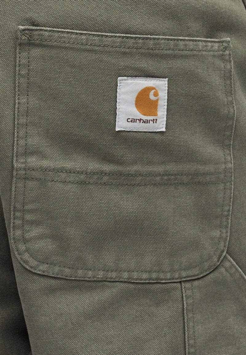 Gros plan sur une poche en tissu vert olive avec un écusson logo Carhartt blanc cousu près du centre supérieur.