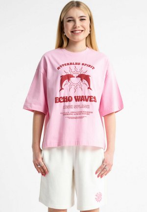 Młoda kobieta uśmiechająca się, ubrana w luźny różowy T-shirt z czerwonym nadrukiem delfina i słońca oraz białe krótkie spodenki sięgające do kolan z małym różowym wzorem.