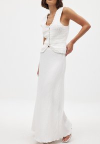 Valge heegeldatud topp, millel on volangiga kaelus, koos pika, tekstureeritud valge seelikuga. Komplektil on vormiv siluett ja nööbiklapp.