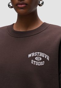 Felpa marrone con collo rotondo, presenta un logo ricamato bianco che legge "WRSTBHVRS STUDIO" sul petto. Tessuto morbido.