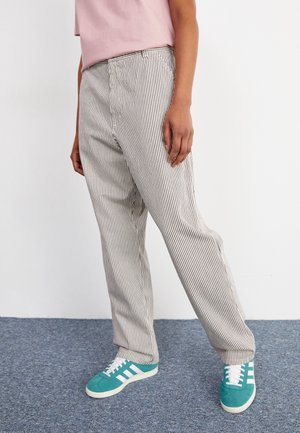 Osoba nosząca czarno-białe spodnie w pionowe paski, różową koszulę i turkusowe trampki, stojąca na niebieskim dywanie.