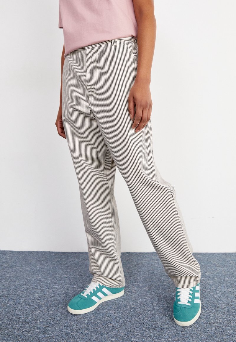 Personne portant un pantalon à rayures verticales noir et blanc, une chemise rose et des baskets bleu sarcelle, debout sur un tapis bleu.