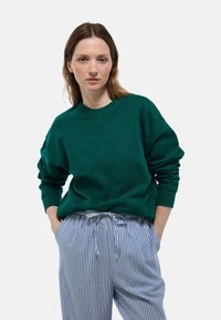 Sweatshirt vert en tissu doux avec un col rond, épaules tombantes et poignets côtelés, associé à un pantalon rayé bleu et blanc.