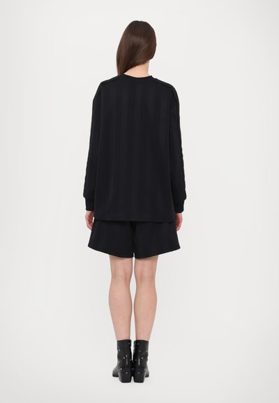 Black oversized long-sleeve top with vertical stripes, paired with black shorts. Model wears ankle boots with decorative hardware.
