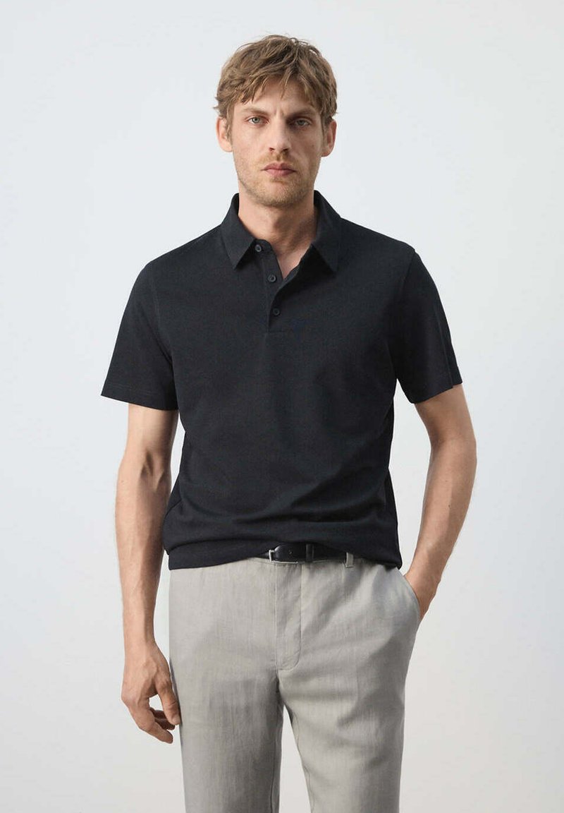 Homme aux cheveux châtain clair portant un polo noir à manches courtes et un pantalon gris clair, debout devant un fond uni.
