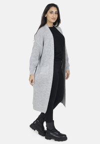 Lange grijze cardigan van chunky breiwerk, met een open voorkant, geribde mouwen en zijzakken, gedragen over een zwart topje en strakke broek.