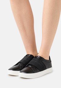 Černé kožené slip-on boty s bílou gumovou podrážkou a elastickými pásky s logem přes přední část. Hladký povrch s minimálními detaily.