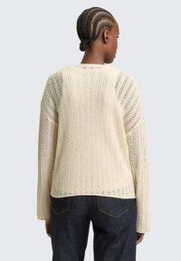 Persoon met gevlochten haar, gekleed in een crèmekleurige gebreide trui met een kantpatroon en donkerblauwe spijkerbroek, van achteren gezien.