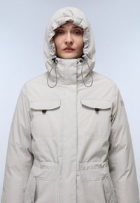 Chaqueta con capucha de color gris claro, hecha de una tela suave y texturizada. Cuenta con dos bolsillos en el pecho, cordones ajustables en la capucha y una cremallera frontal.