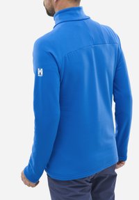 Mann trägt eine leuchtend blaue Fleecejacke mit langen Ärmeln und hohem Kragen sowie dem Millet-Logo auf dem linken Ärmel, abgewandt.