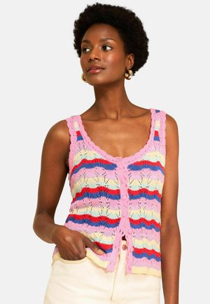 Kvinde med kort krøllet hår iført guld øreringe i hoop-stil og en lyserød strikket top med pastelfarvede striber og knapper, hånd i lommen.