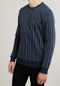 Námořnický svetr s kulatým výstřihem a vertikálními bílými pruhy, s žebrovanými manžetami a lemem, na kterém je malá černá značka na hrudi. Texturovaná tkanina.
