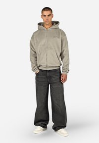 Pelēks fliša džemperis ar rāvējslēdzēju, priekšējā kabata un ribota aproces, kombinēts ar brīvi piegulošām tumšām džinsa biksēm un baltām sporta kurpēm.