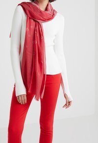 Écharpe rouge à texture légère et transparente avec un motif discret de logo, drapée sur un haut à manches longues blanc, associée à un pantalon rouge.