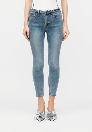 Mujer con vaqueros ajustados de color azul claro y tacones plateados de punta puntiaguda con grandes lazos, posando contra un fondo liso.