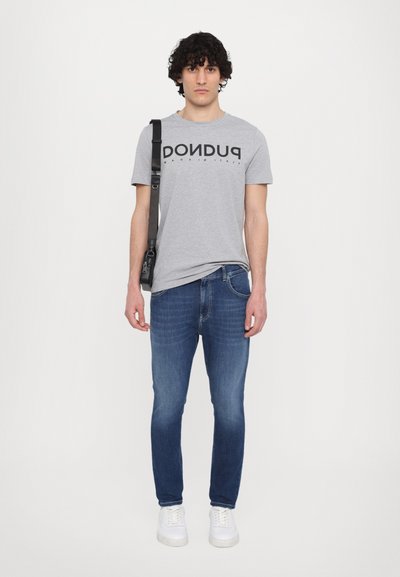 Grå bomull T-skjorte med svart tekst, kombinert med blå denimjeans. Modellen har på seg hvite joggesko og bærer en svart skulderveske.