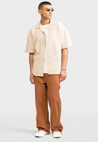 Light beige short-sleeve button-up shirt with a geometric pattern, paired with loose brown pants featuring pockets and white sneakers.