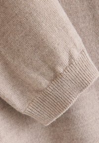 Gestrickter beiger Pulloverstoff mit einer strukturierten Oberfläche. Nahaufnahme des Bündchens zeigt gerippte Details und eine weiche, glatte Textur.