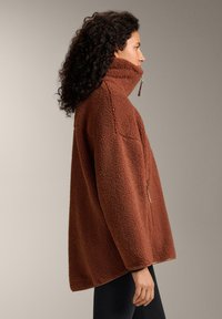 Brown fleece pullover with a high collar, featuring a zip closure and a loose fit. Textured fabric adds warmth and dimension.
